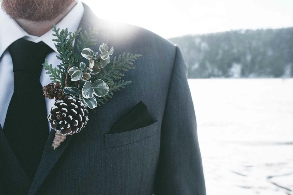 A close-up of a boutonniere pinned on a suit jacket featuring green sprigs and a frosted pinecone