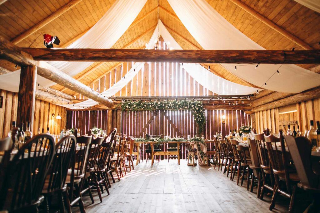 A rustic wedding inside a barn with twinkle lights and draped fabric hanging from the building peak. Wooden chairs and long tables are set up beneath