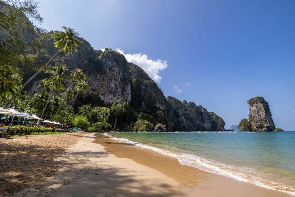 A view of Ao Nang Beach in Thailand during the day with water receding from the sandy shores and palm trees and mountain cliffs in the background