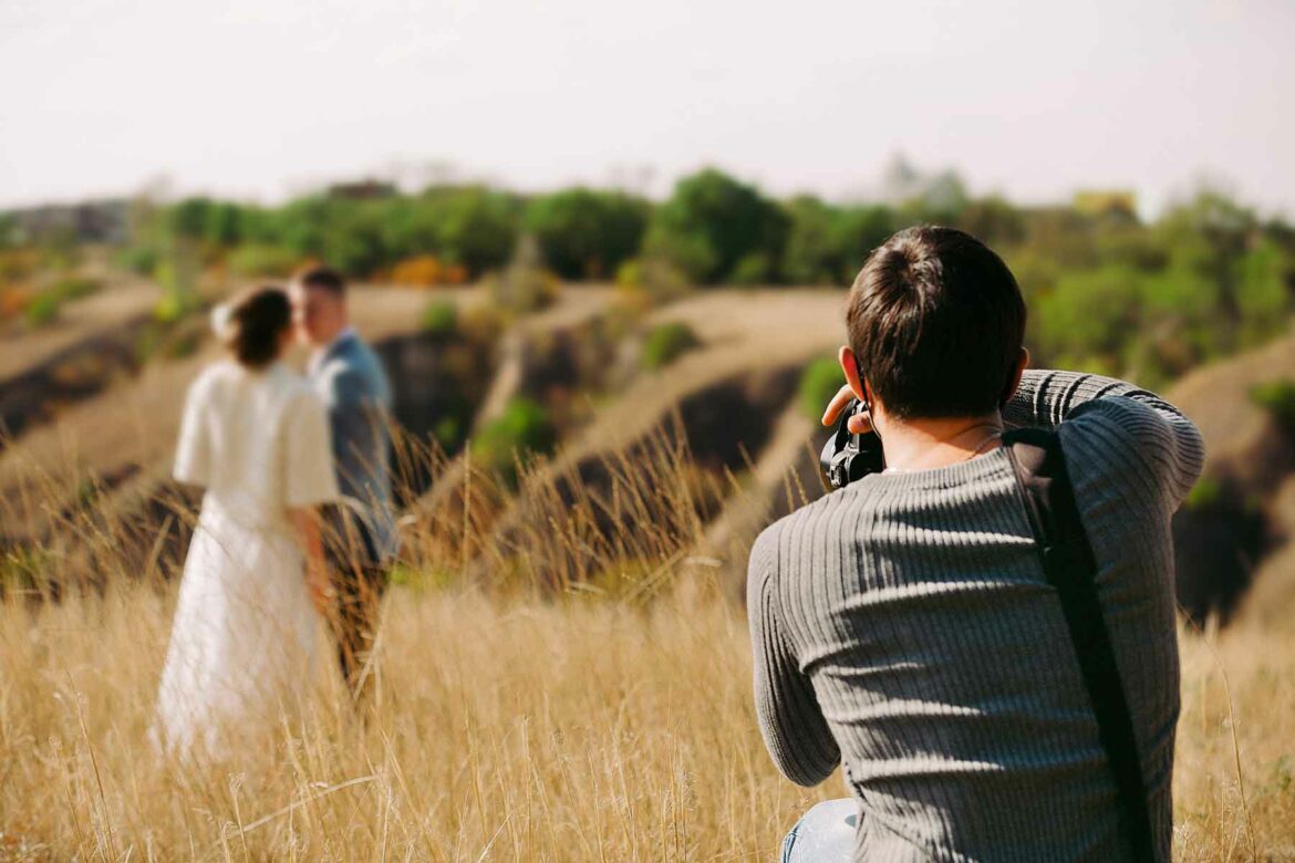 An engagement photoshoot taking place in a meadow with a couple embracing in the background and an engagement photographer in the foreground