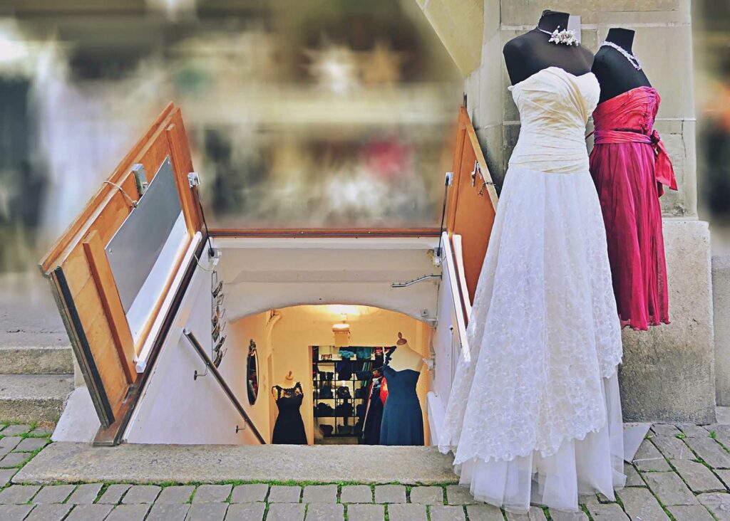 A wedding dress and bridesmaid dress on mannequins in front of a doors opening to a below-ground bridal salon down a flight of stairs