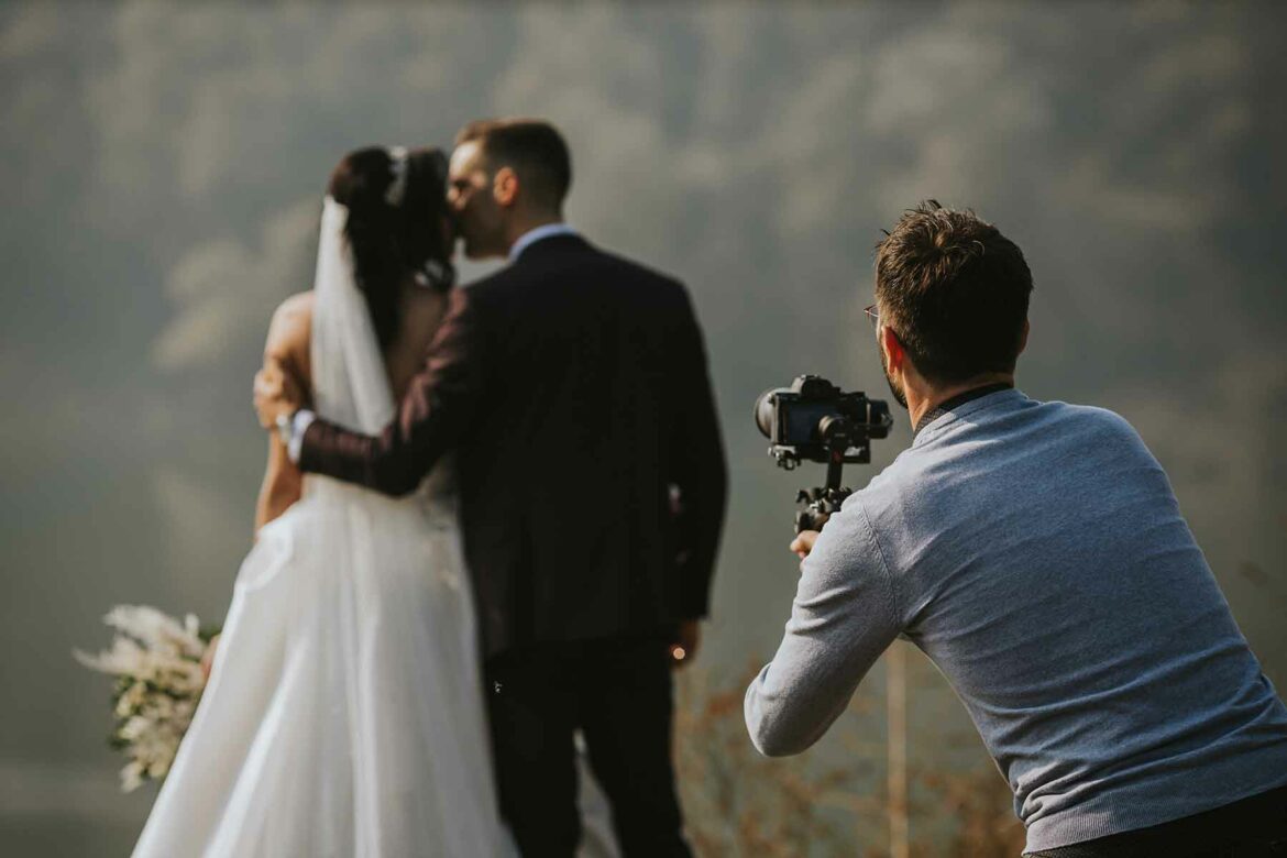 A wedding photographer takes a photo of a bride and groom from behind while standing on a hillside