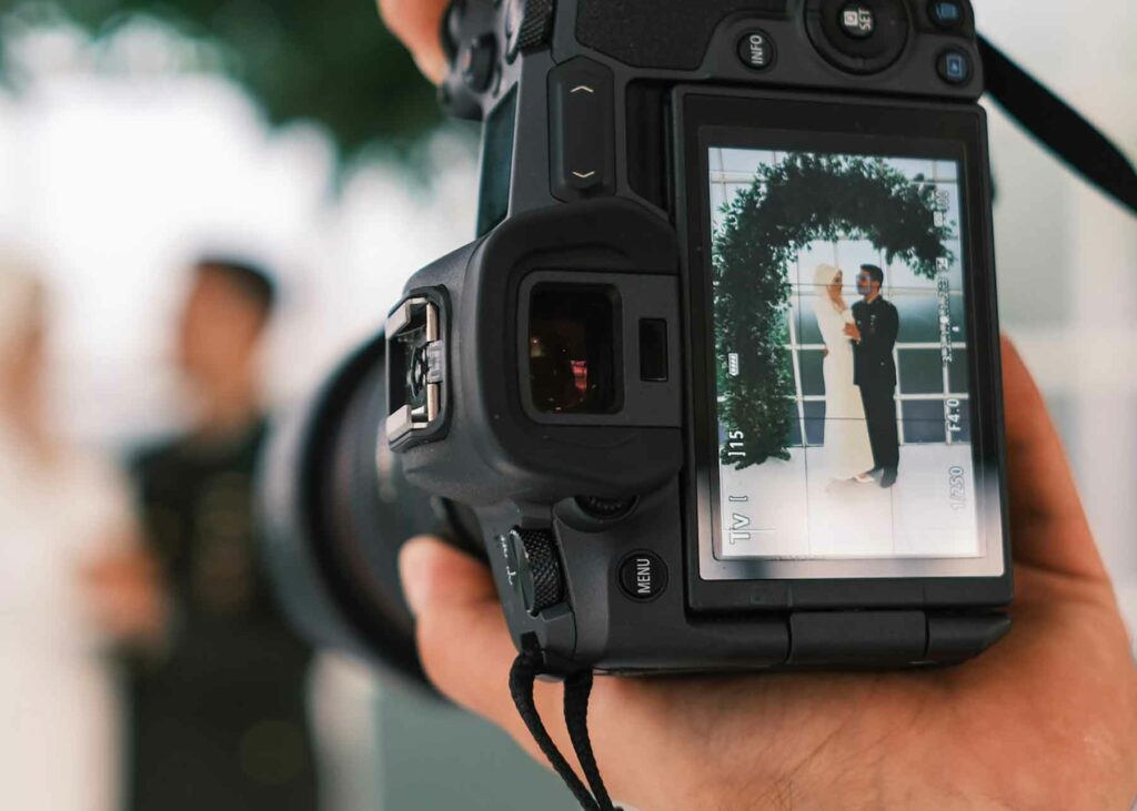A close-up of a DSLR camera held by a wedding photographer that shows a bride and groom under a green arch in the viewfinder