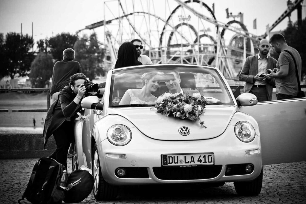 Black and white photo of wedding photographer capturing bride and groom as they leave in the getaway car with an amusement park in the background