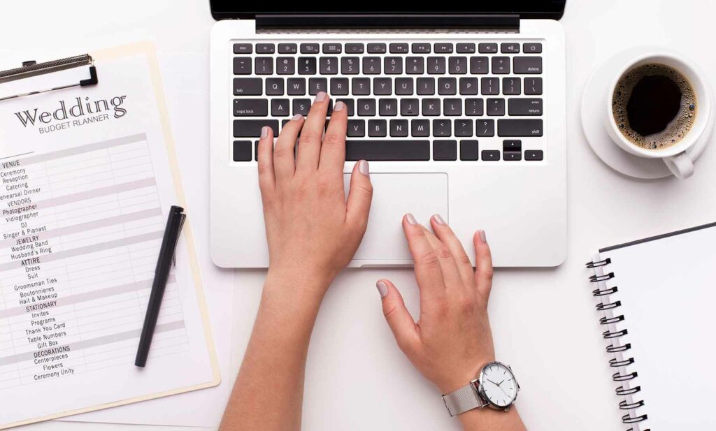 An image of a set of hands at a laptop with a checklist that reads "wedding budget planner to the left and a cup of coffee to the right