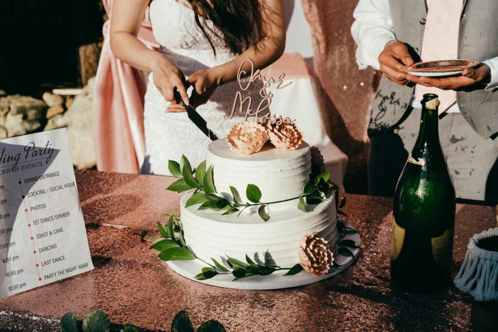 A bride and groom cutting their wedding cake. The cake is two-tiered and embellished with greenery and pale pink flowers. 