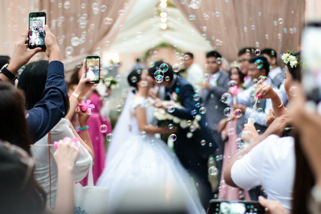 A bride and groom kissing in the background of a wedding surrounded by bubbles while guests take photos on their phones