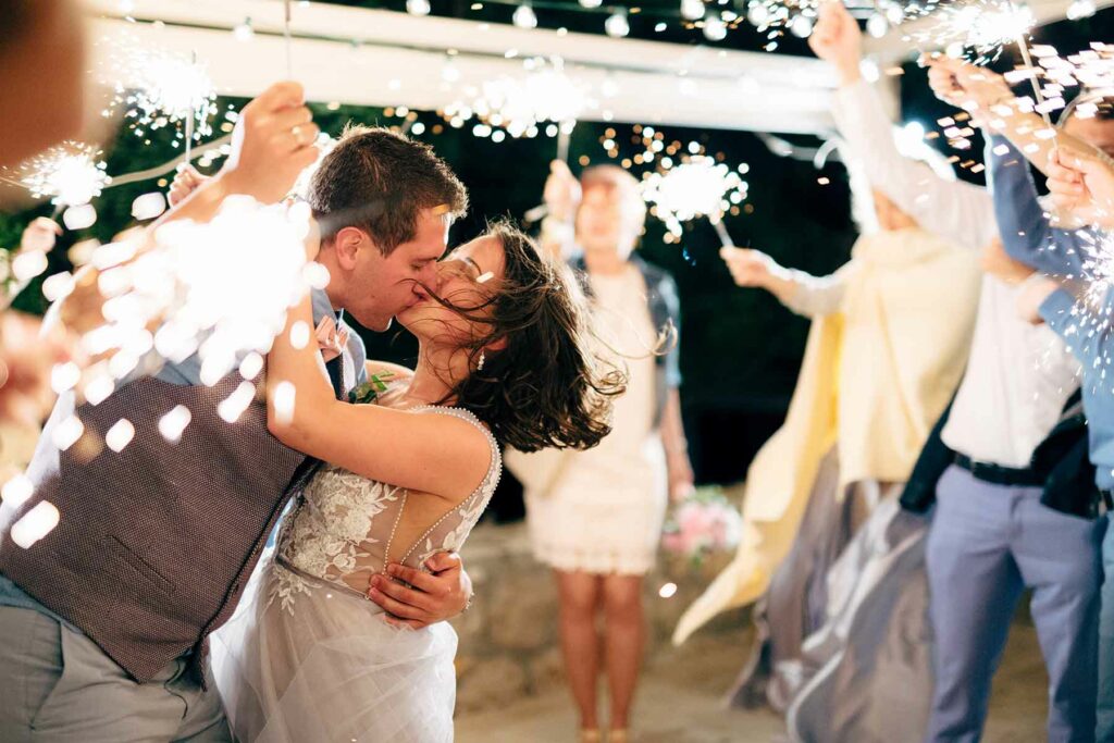A couple in wedding attire kissing and embracing while wedding guests look on and hold sparklers