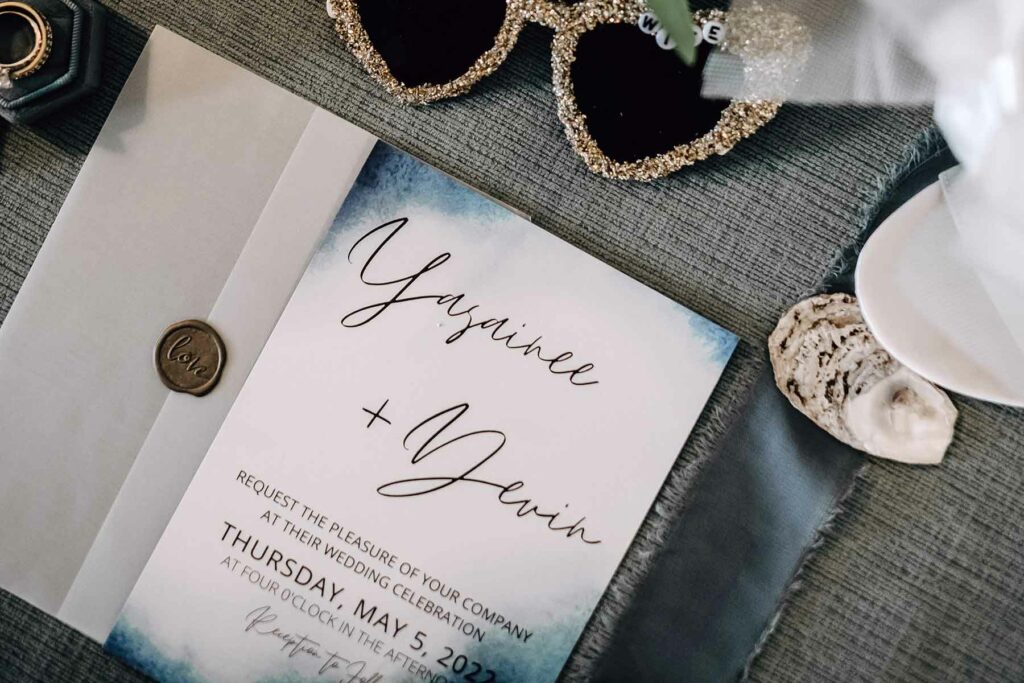 A wedding invitation with watercolor edges  surrounded by sunglasses, wedding rings, and a seashell