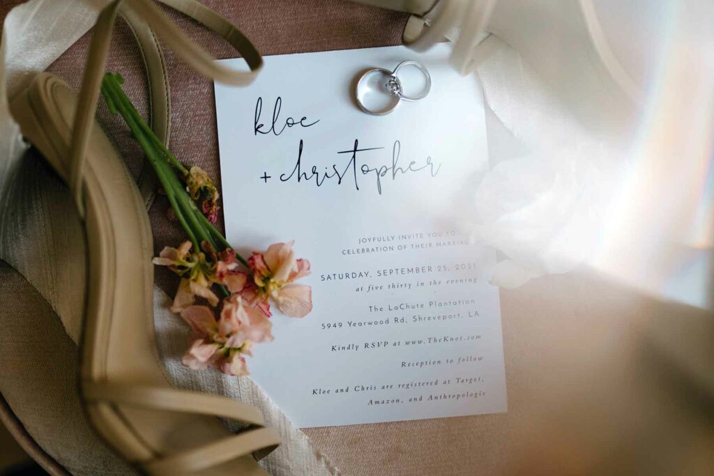 A wedding invitation with two rings laid on top of it, surrounded by flowers, a pair of heels and slightly lit up from the glow of sunlight