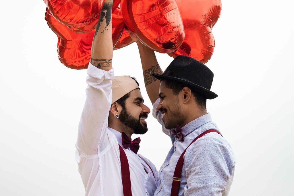 Two men looking into each other's eyes wearing matching suspenders and holding red heart balloons