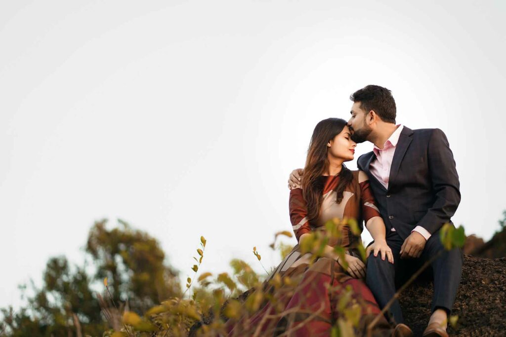What to Wear in Engagement Photos: A woman wearing a maroon dress being kissed on the forehead by a man in a dark brown suit