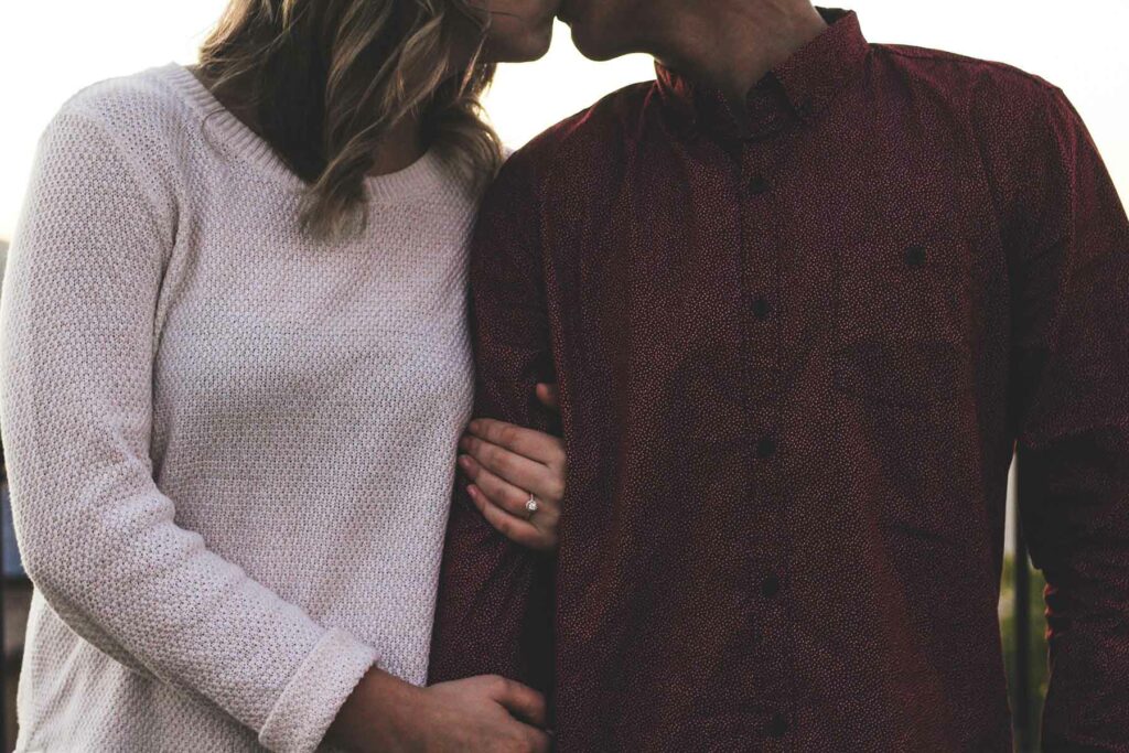 What to Wear in Engagement Photos: A woman in a beige sweater and engagement ring holding the arm of a man in a patterned shirt