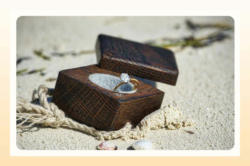 A diamond ring in a wooden box buried partway in the sand