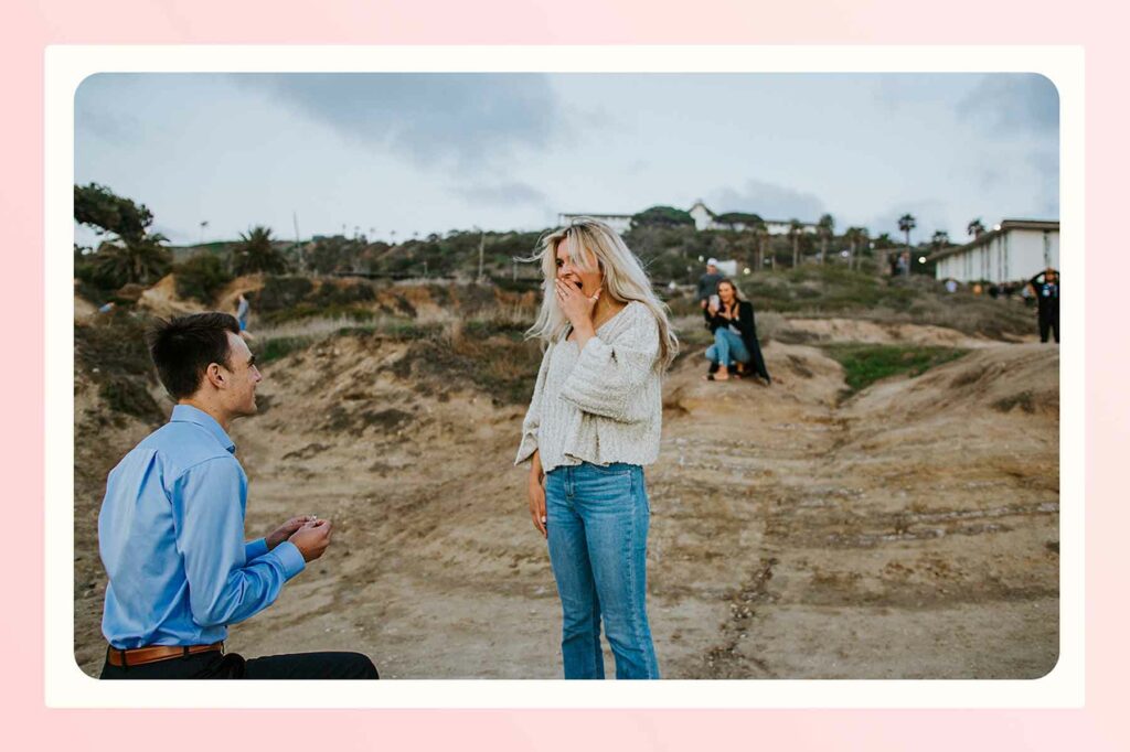 A beach proposal with friends and family taking pictures of the couple from the cliffs