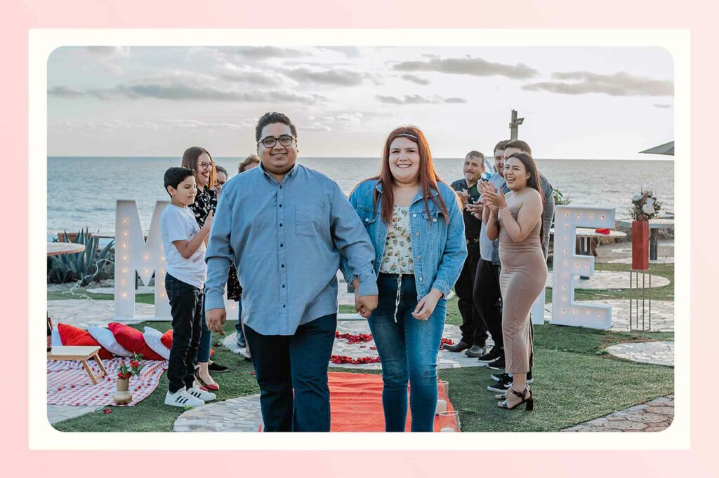 A smiling couple holding hands walking away from their beach proposal with friends and family clapping for them in the background