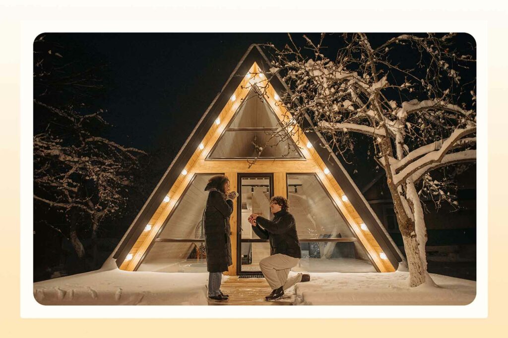 A man on one knee in the snow in front of an A-frame lit up in the night sky, about to propose