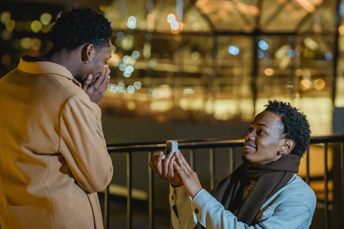 A person kneeling and holding a ring box out during a proposal with city lights at night in the background