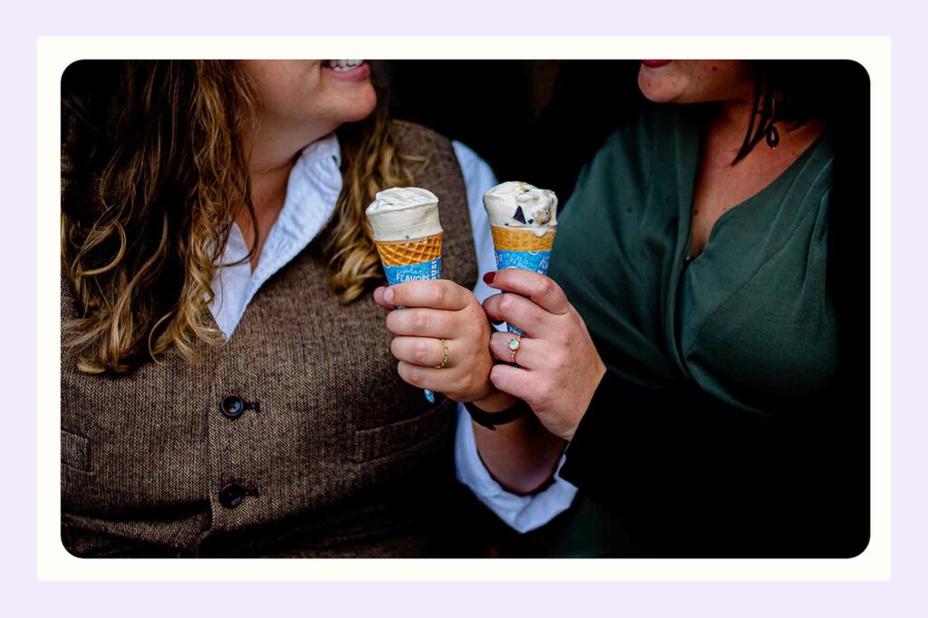 A close-up of two people holding ice cream cones. One woman is wearing an engagement ring