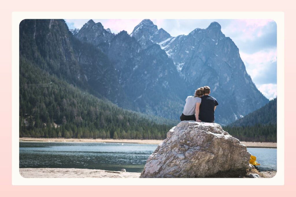 A couple sits with their backs to the camera atop a large rock by a lake surrounded by large mountains