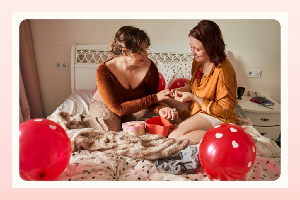 Female couple sitting in bed, surrounded by red balloons, while one woman slips a ring on the finger of the other woman