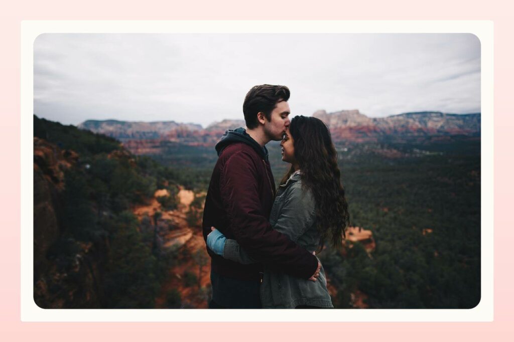Couple standing in front of beautiful outdoor landscape hugging while man kisses woman's forehead