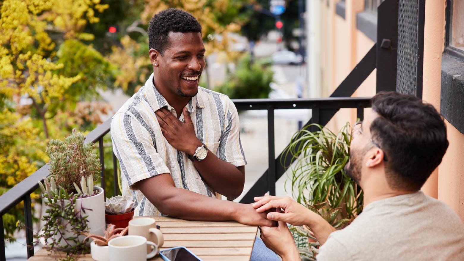 Man sitting at outdoor patio table surprised by his partner kneeling down and proposing while holding his hand
