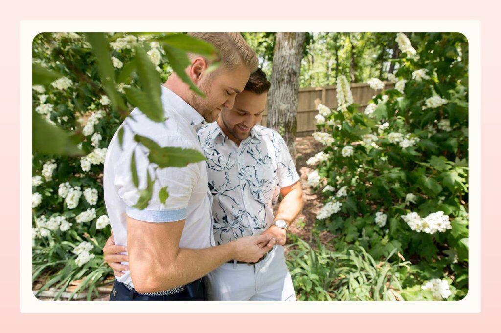 Male couple stands in garden holding hands and both looking at the engagement ring on one of their hands