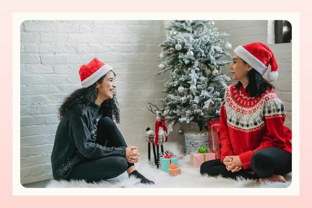 Two women with Santa hats on sit beside a Christmas tree while looking at each other