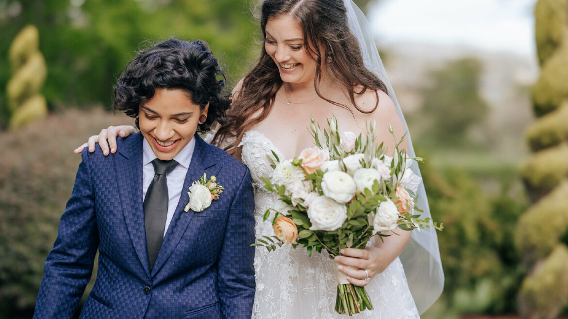 A lesbian wedding couple, one in a gender neutral wedding suit and the other in a white dress with bouquet, walk together with smiles