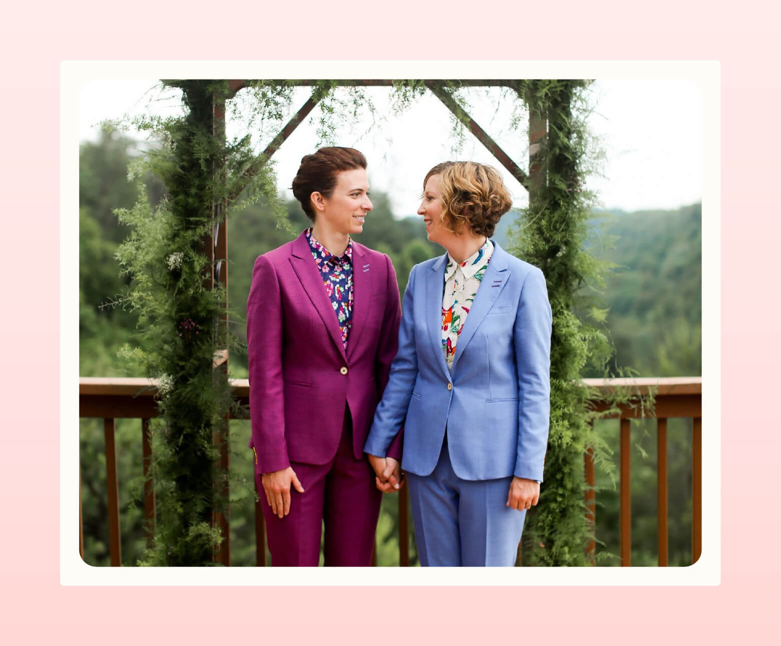 A couple in complimentary suits, one fuchsia and the other pale blue, from Sharpe Suiting holds hands and look at each other standing in front of greenery arch
