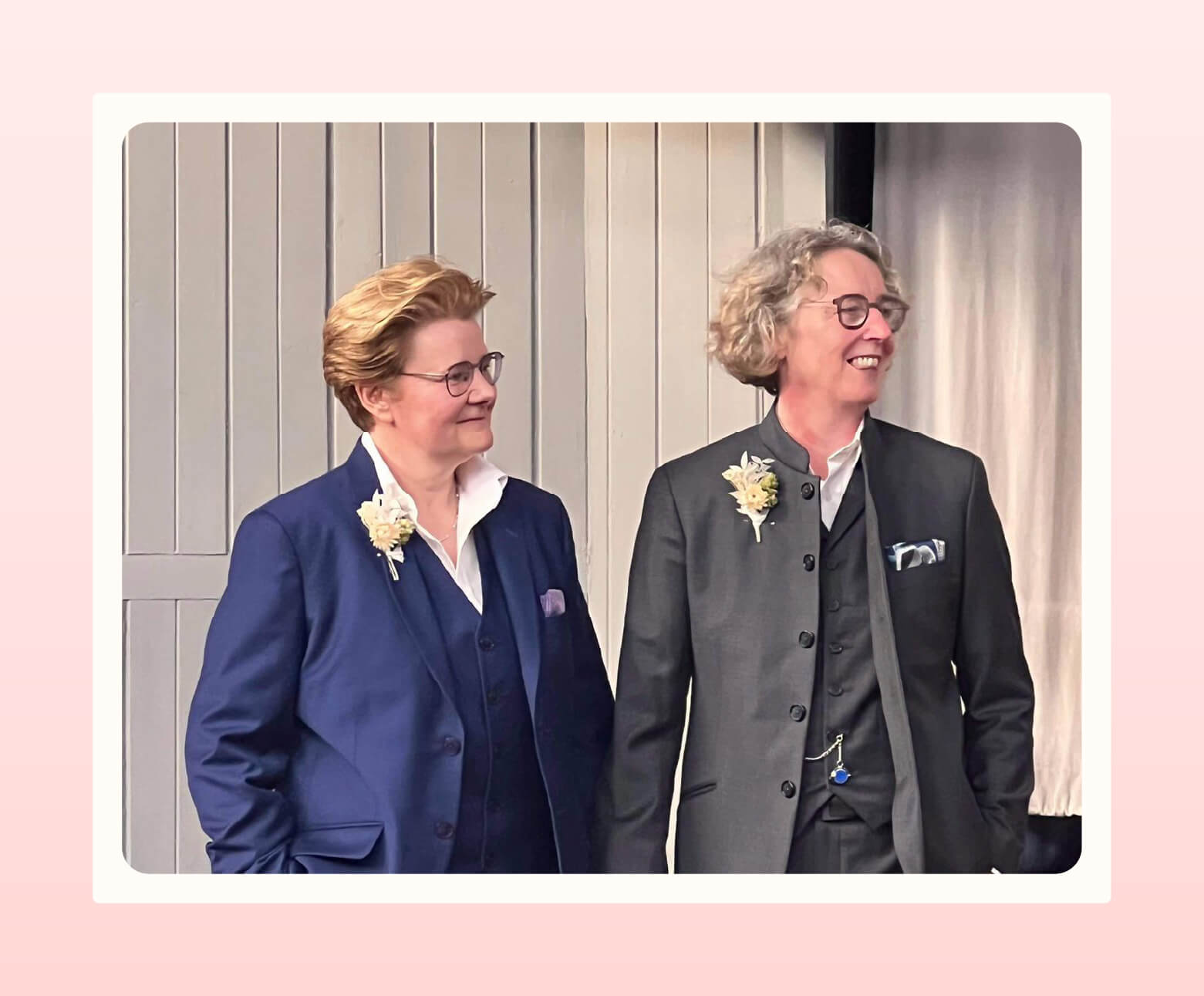 A couple holding hands, one wearing a blue suit and the other wearing a grey suit, both from The Butch Clothing Company, look to their left with smiles on their faces