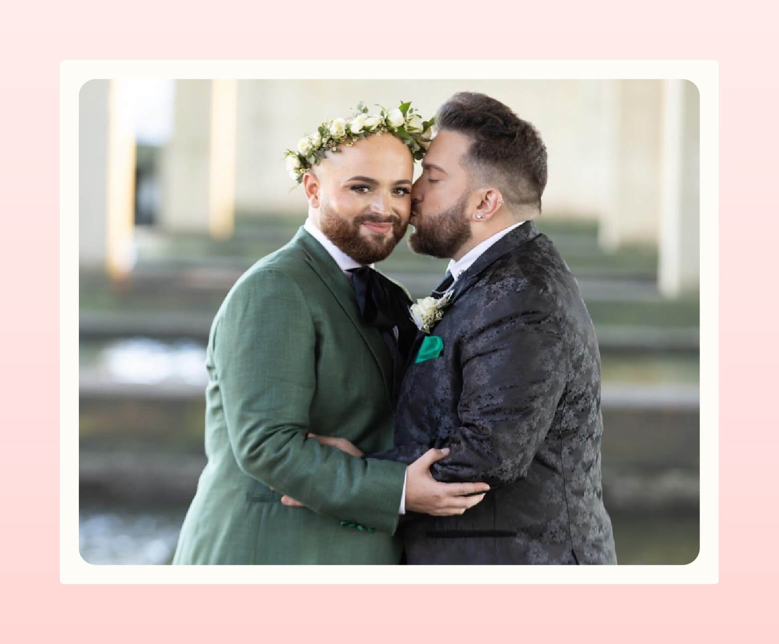 Wedding couple with short beards pose, one looking to camera wearing a green suit jacket and a flower crown while the other kisses their cheek and wears a black textured suit jacket with boutonniere
