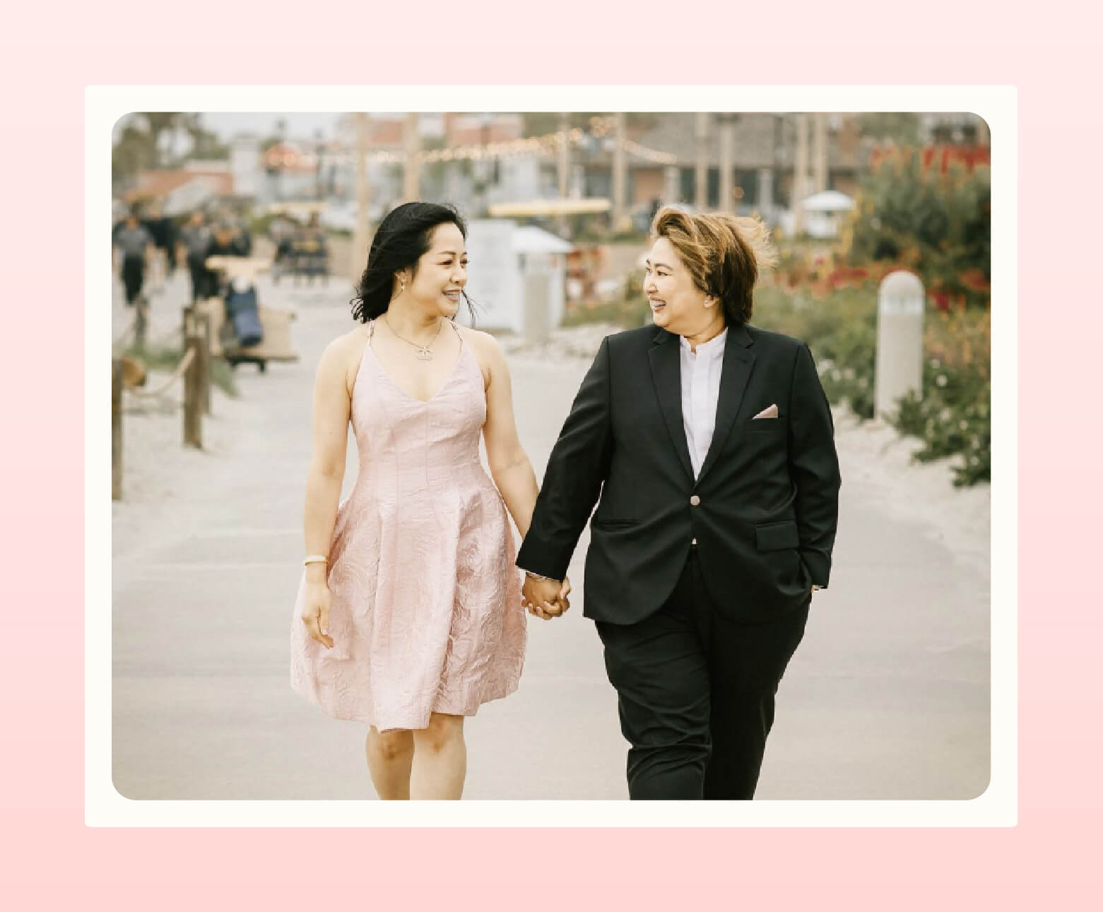 Couple walks holding hands, one wearing black suit from Thúy Custom Clothier and the other wearing a pale pink knee-length dress