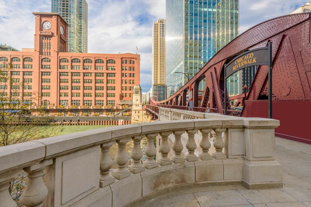 Chicago Riverwalk sign with a bridge, stone walkway and buildings in the background