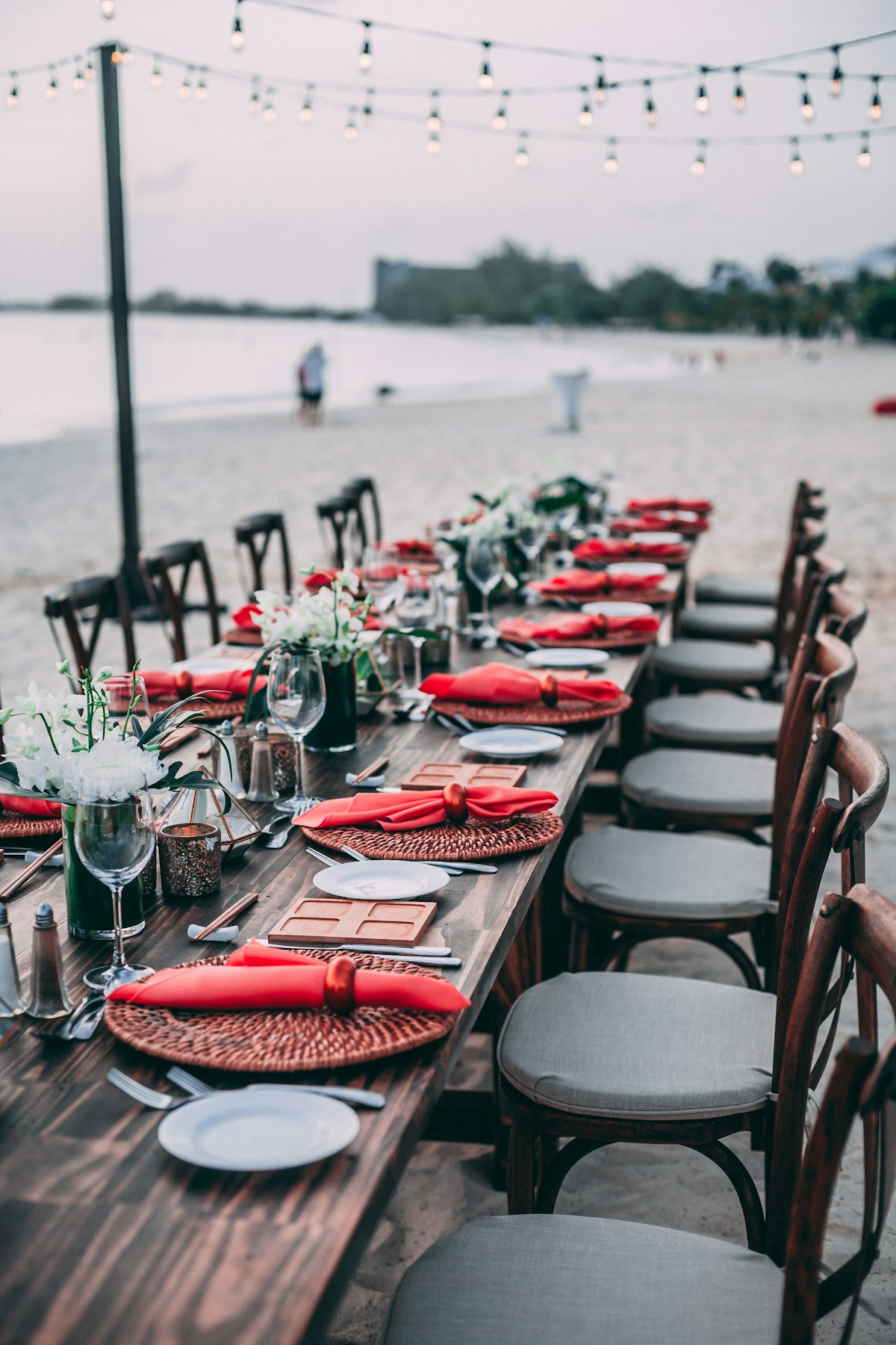An elegant destination wedding reception table set up on a beach with string lights and ocean views at sunset